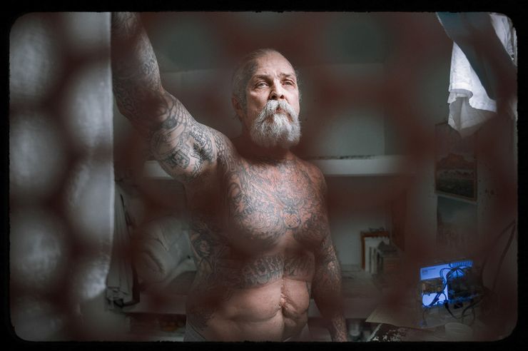 A photo shows Craig Canary, a White man with a white beard and tattoos visible on his upper body, standing in a solitary confinement cell. Visible in his cell are a bedroll, a few books and some art on his wall. In the foreground of the photo is a door with holes, through which you can see Canary.     