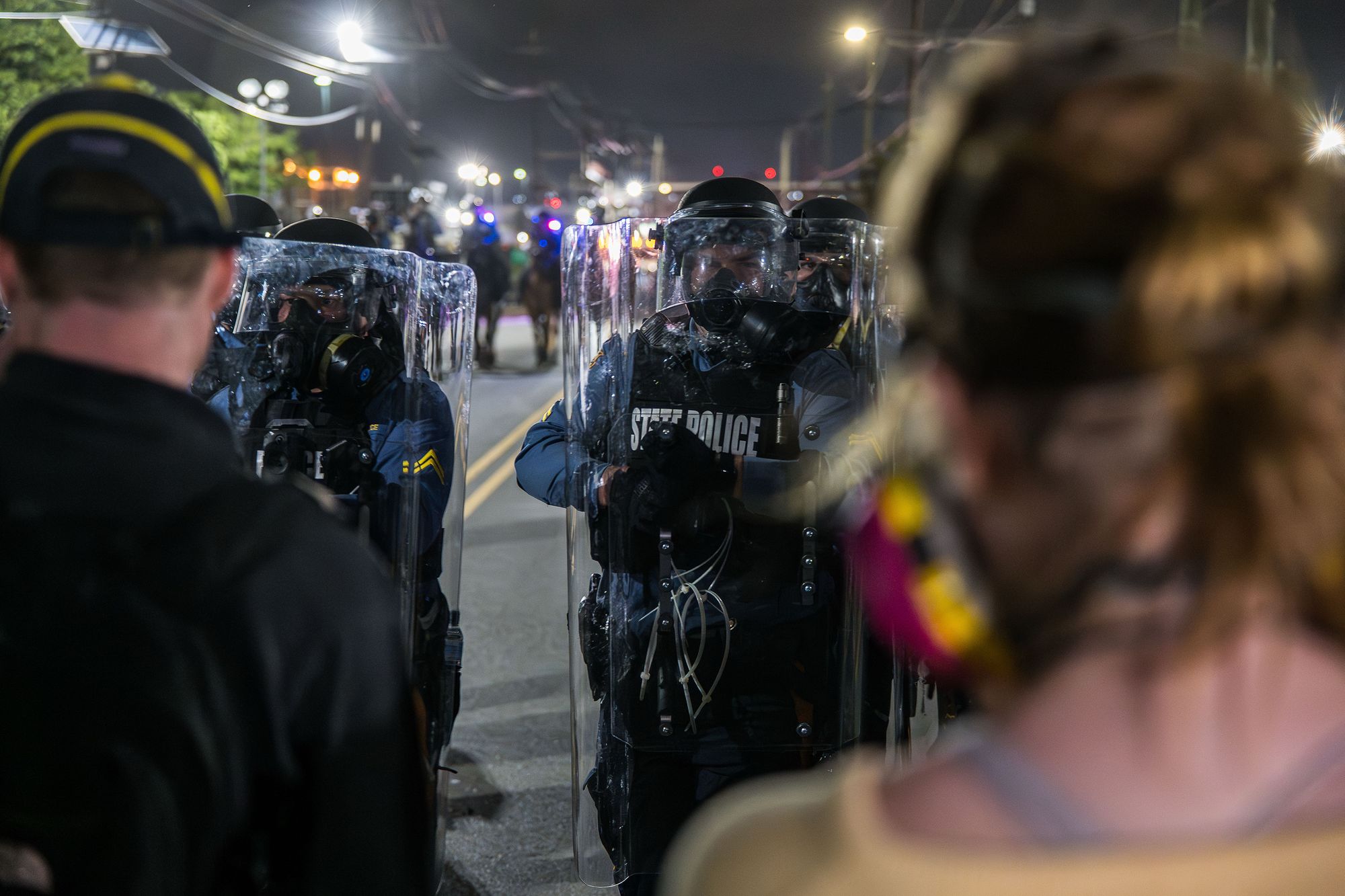 A photo shows state troopers in riot gear at night facing two protesters whose backs are to the camera.  