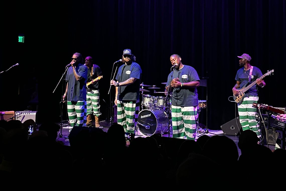 A photo shows five men performing on a stage, all wearing dark blue T-shirts and green-and-white striped pants. Four of the five men are Black, and two of them are holding instruments.