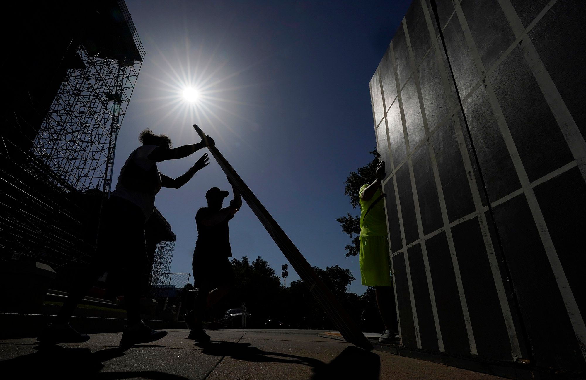 A photo shows the sun beaming above silhouettes of two people holding a board while another person constructs a wall of a makeshift prison cell. 