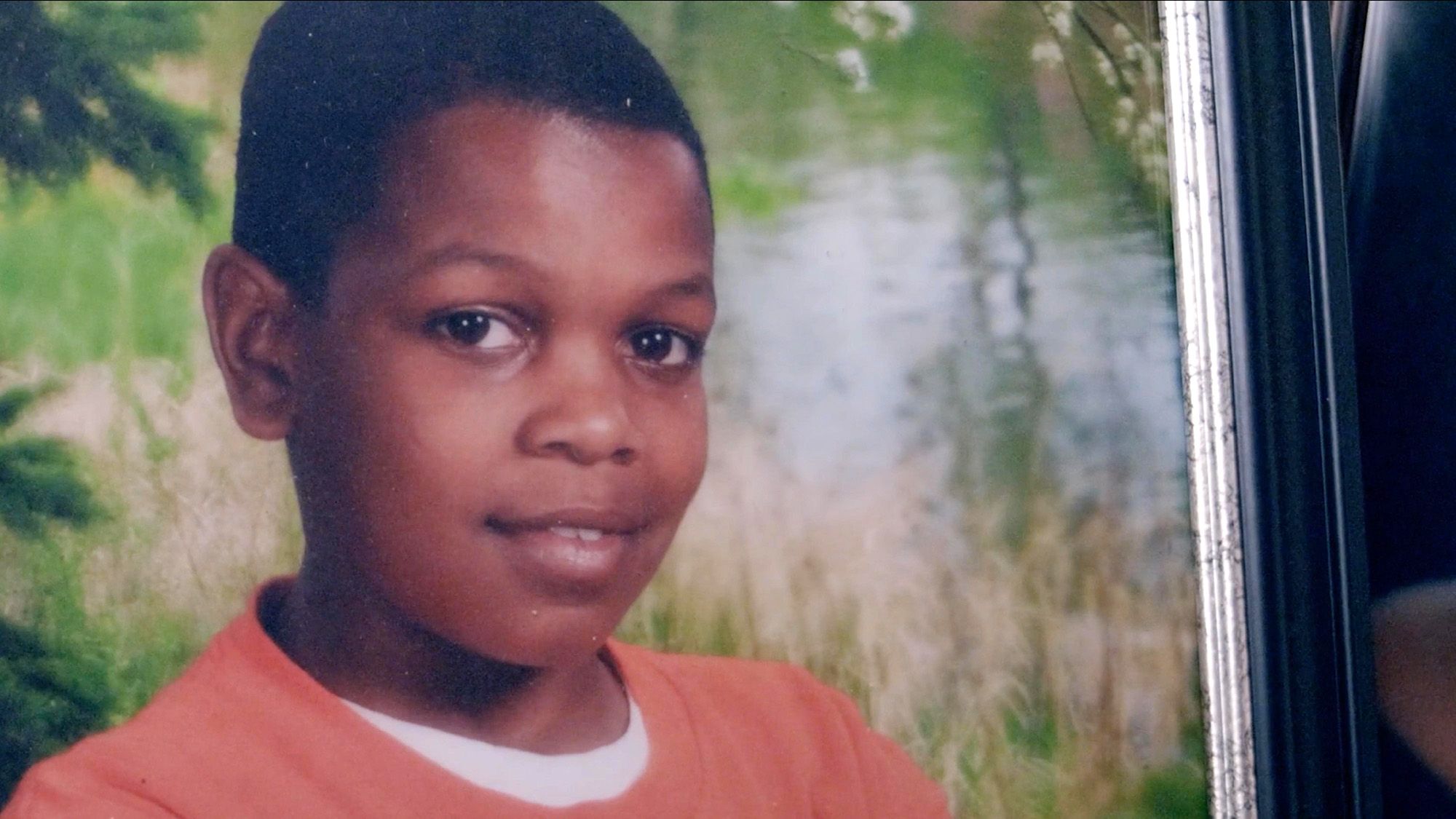 A photo shows a Black boy in an orange shirt posing for a school portrait. 