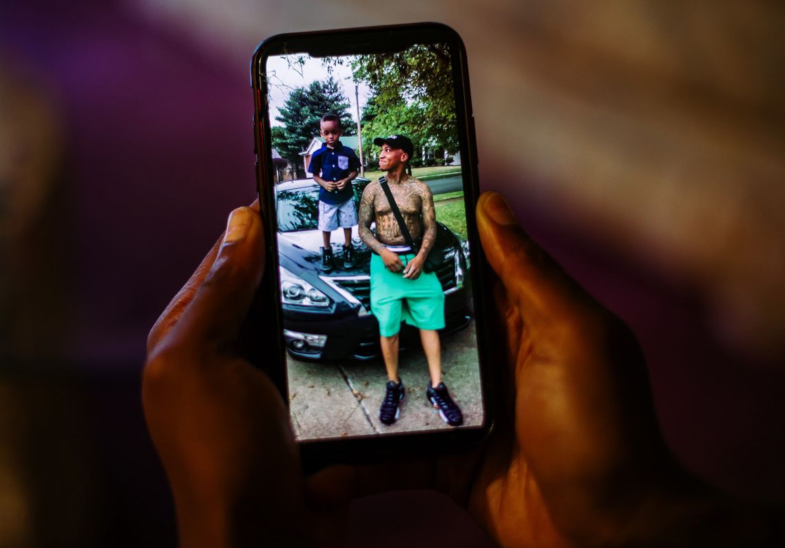 A pair of hands holds a phone, displaying an image of a Black man looking at his young son as he stands on the hood of a car. 