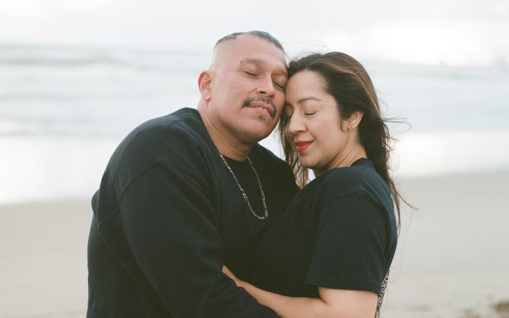 Miguel, a man with medium-toned skin, short hair and a mustache, hugs his wife, Silvia, a woman with medium-toned skin and long brown hair, on the beach.  