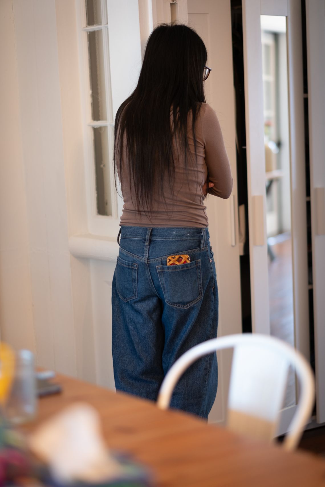 Una mujer se vuelve lejos de la cámara. Tiene cabello largo, castaño oscuro. Viste una blusa de manga larga café claro y un pantalón de mezclilla. Está de pie en una habitación con una mesa de madera detrás de ella. 
