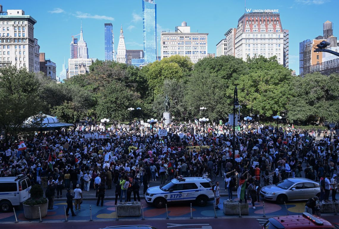 A photograph shows a large group of protesters in a clearing, with trees and skyscrapers behind them. Police cars are parked near the crowd. 