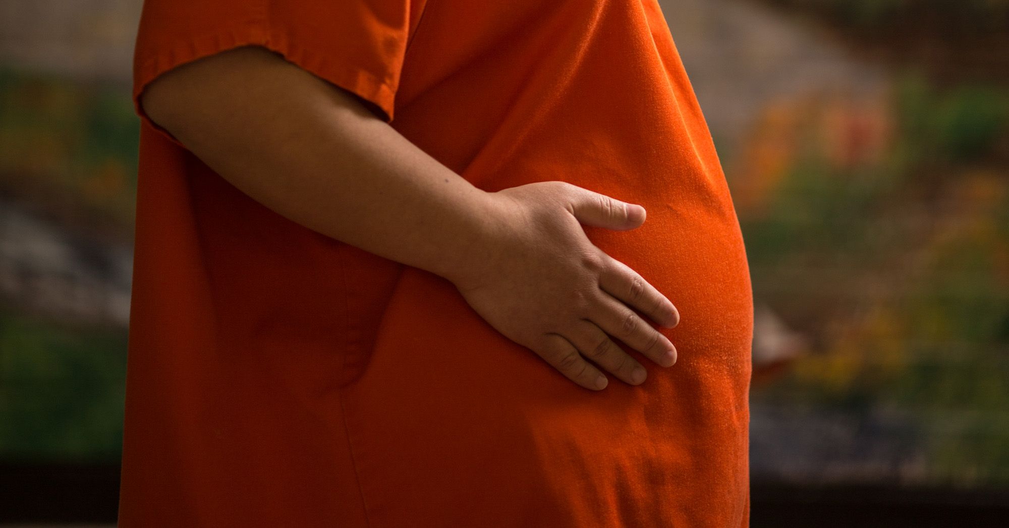 A pregnant woman wearing an orange prison uniform places her hand on her belly. 