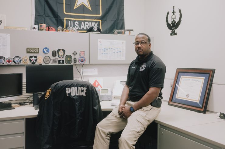 A Black man with glasses, wearing a Black polo and khakis with handcuffs visible from his holster, sits at his cubicle, next to a chair with a jacket draped over it that says POLICE. Badges adorn the cabinet in front of the chair, and a U.S. Army banner hangs above.