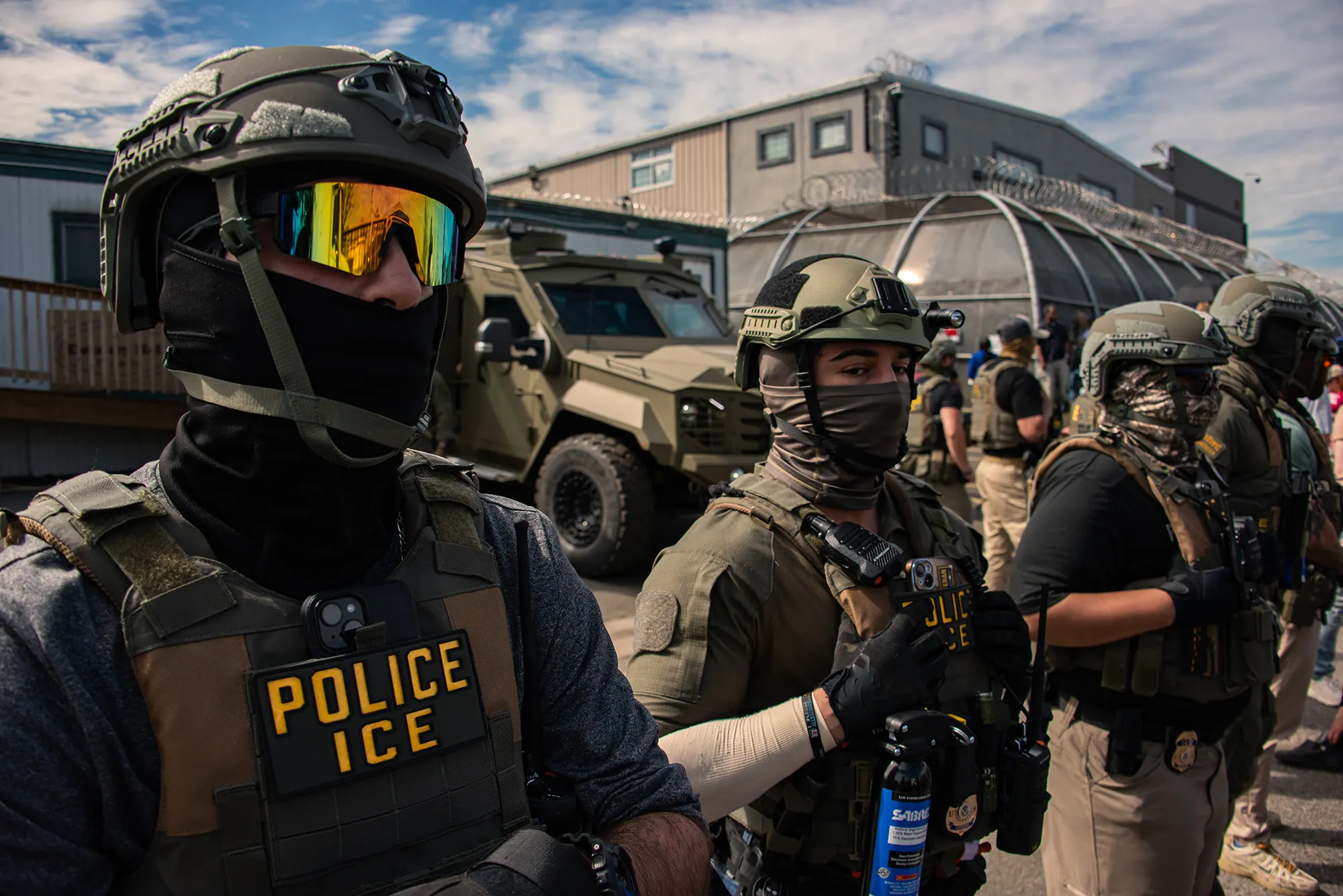 A photo shows several Immigration and Customs Enforcement agents, wearing helmets, masks and vests, standing in front of a military truck and a gray building. 