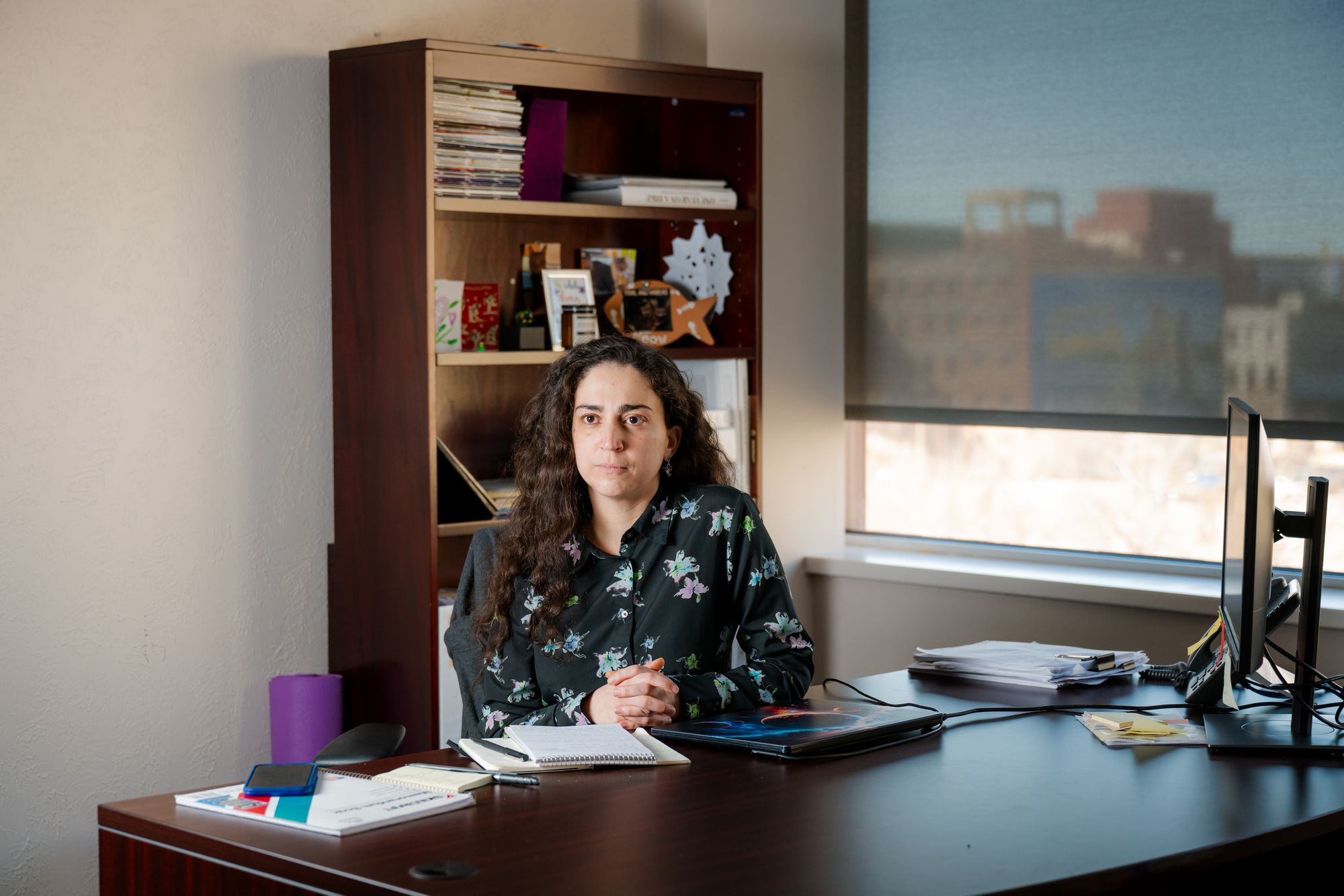A photo shows a White woman with long curly hair, wearing a floral long-sleeved shirt. She is sitting at a desk in an office, with a bookshelf behind her and a window to her left. 