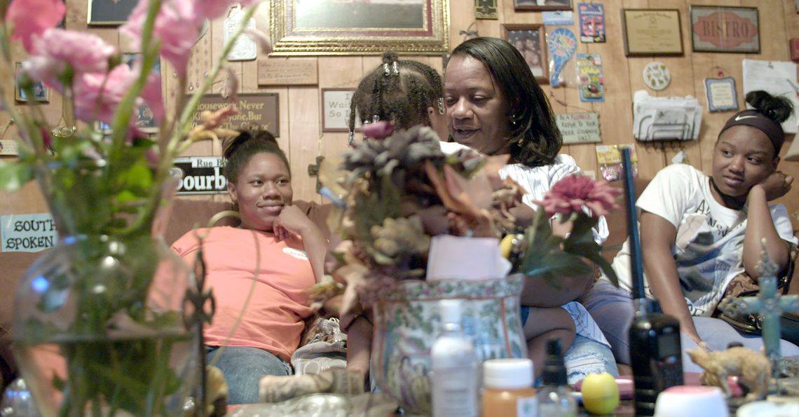 Angela Williams, a Black woman wearing a white T-shirt and blue jeans, holds one of her granddaughters. In the background, her family members sit on a couch.