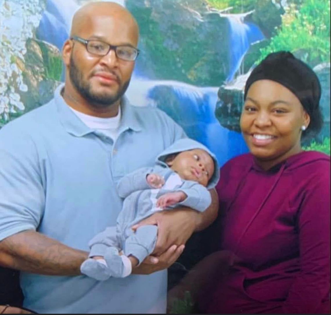 Una mujer Negra con un turbante negro y un suéter magenta sonríe para una foto junto a su padre, quien viste un uniforme gris de prisión. Su padre sostiene a un bebé.