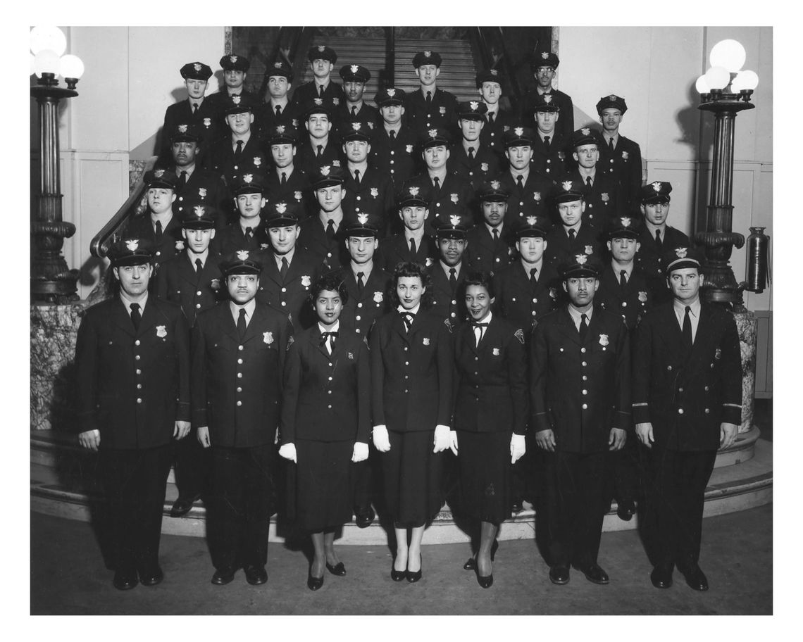 A black-and-white photo shows a group of uniformed police officers, including both men and women, standing in several rows on stairs. The officers are all wearing dark uniforms and caps, and they are posing for a formal group portrait inside a building.