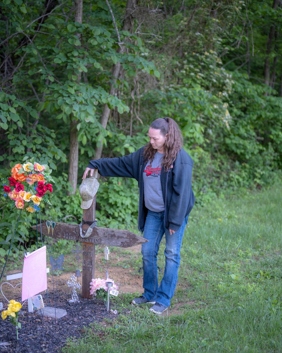 Helphenstine, a White woman with long brown hair in a ponytail, rests her hand on the top of a wooden cross marking the grave of her husband. Flowers adorn the grave.