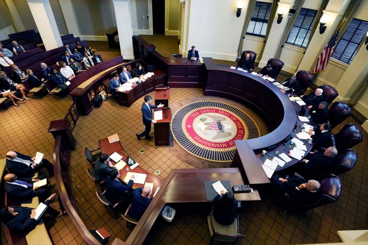 A photo shows an overview of the Mississippi Supreme Court floor. Eight justices sit in large leather chairs at a curved table, while a White man in a suit speaks from a podium near a round rug with the State Seal of Mississippi. People sitting at tables and benches opposite the justices are listening to the speaker.