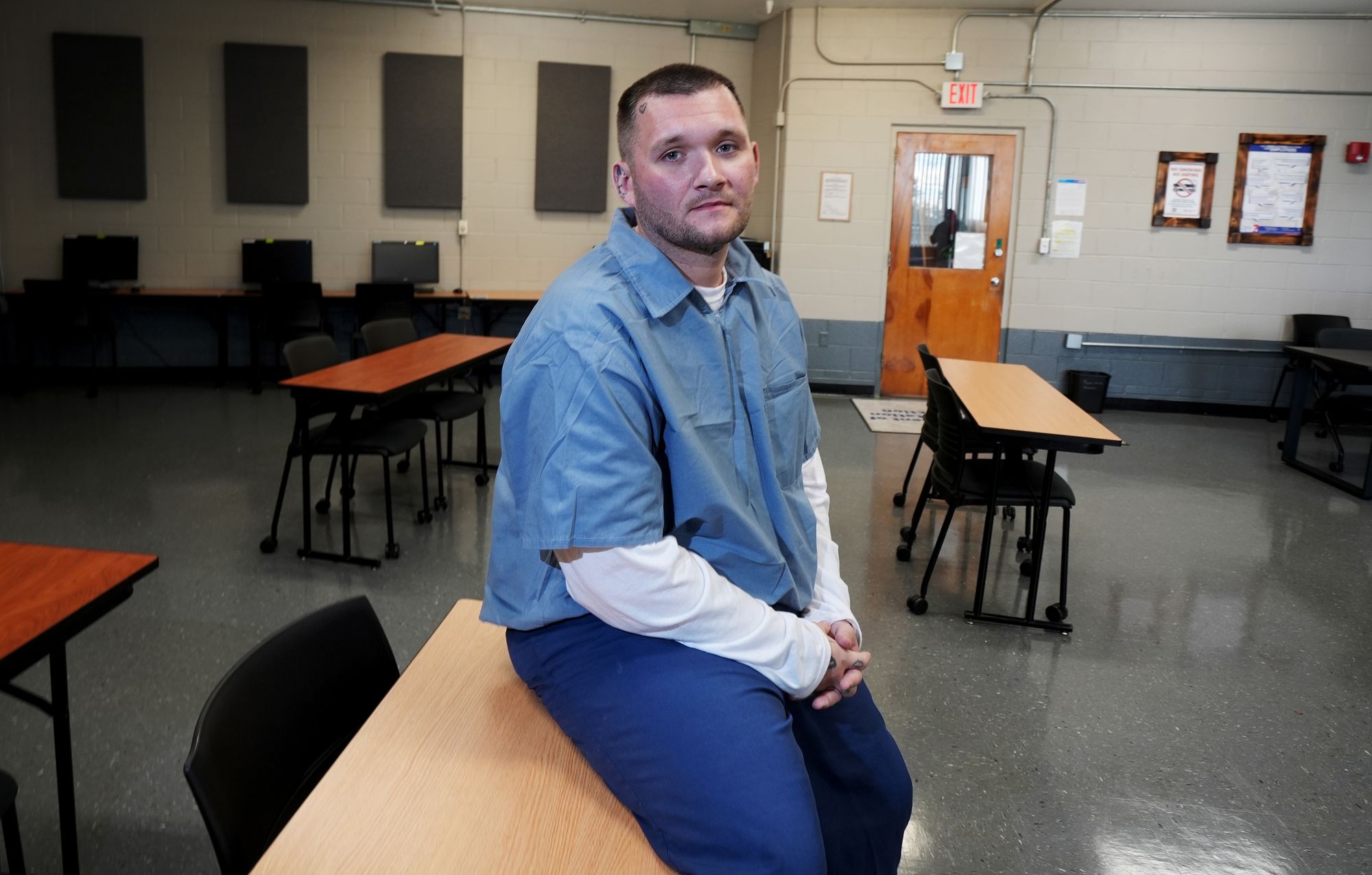 A photo shows Cory Sutphin, a White man wearing a light blue short-sleeved shirt with a white long-sleeved shirt under it, and dark blue pants, which are part of a uniform, sitting on a desk in a room with other desks and chairs in the background. 