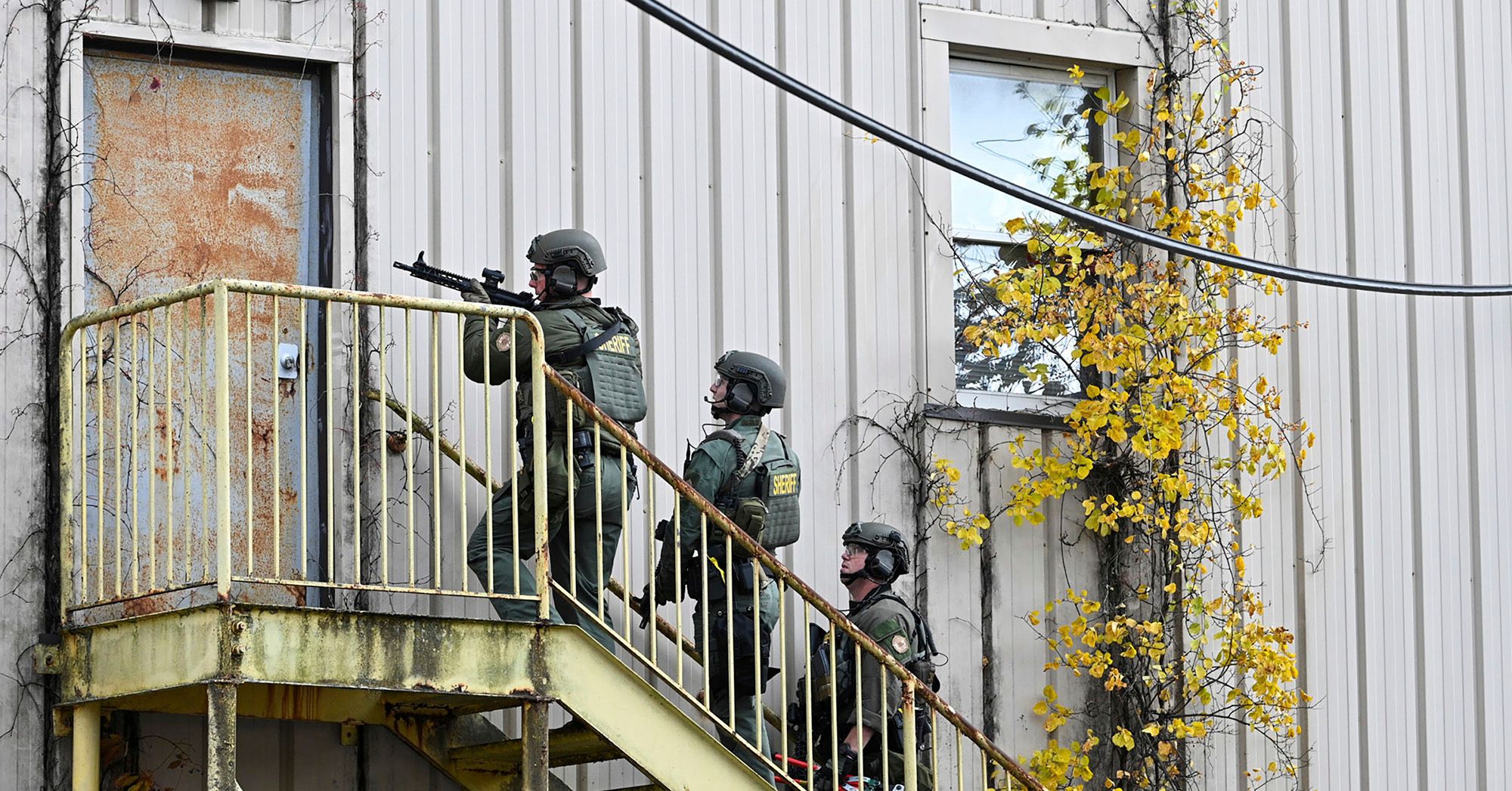 Three SWAT team members, wearing helmets, green uniforms and protective gear with the label "SHERIFF" on their backs, hold assault rifles while standing on a staircase. 