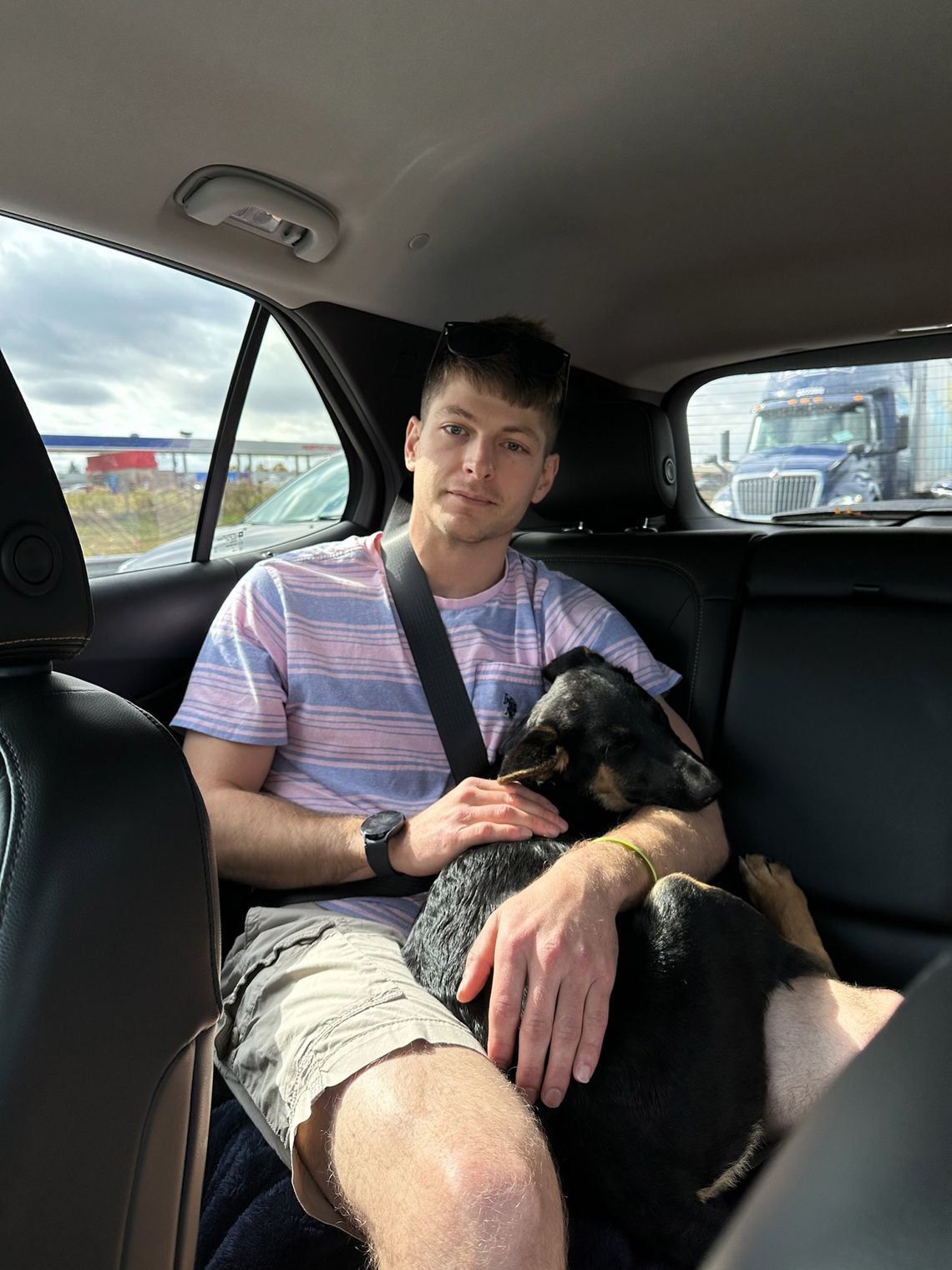 A photo of a young white man wearing a blue-and-pink striped shirt and tan cargo shorts. He is sitting in the back seat of a car holding a black dog.