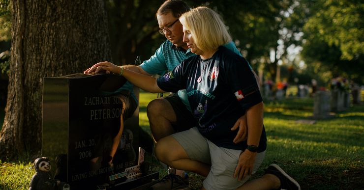 Ronnie Peterson, a man with short brown hair, light-toned skin and wearing a turquoise polo shirt, and Bridget Peterson, a woman with medium-length blonde hair, light-toned skin and wearing a navy blue T-shirt, kneel at the grave of their son, with their hands resting on the gravestone.  