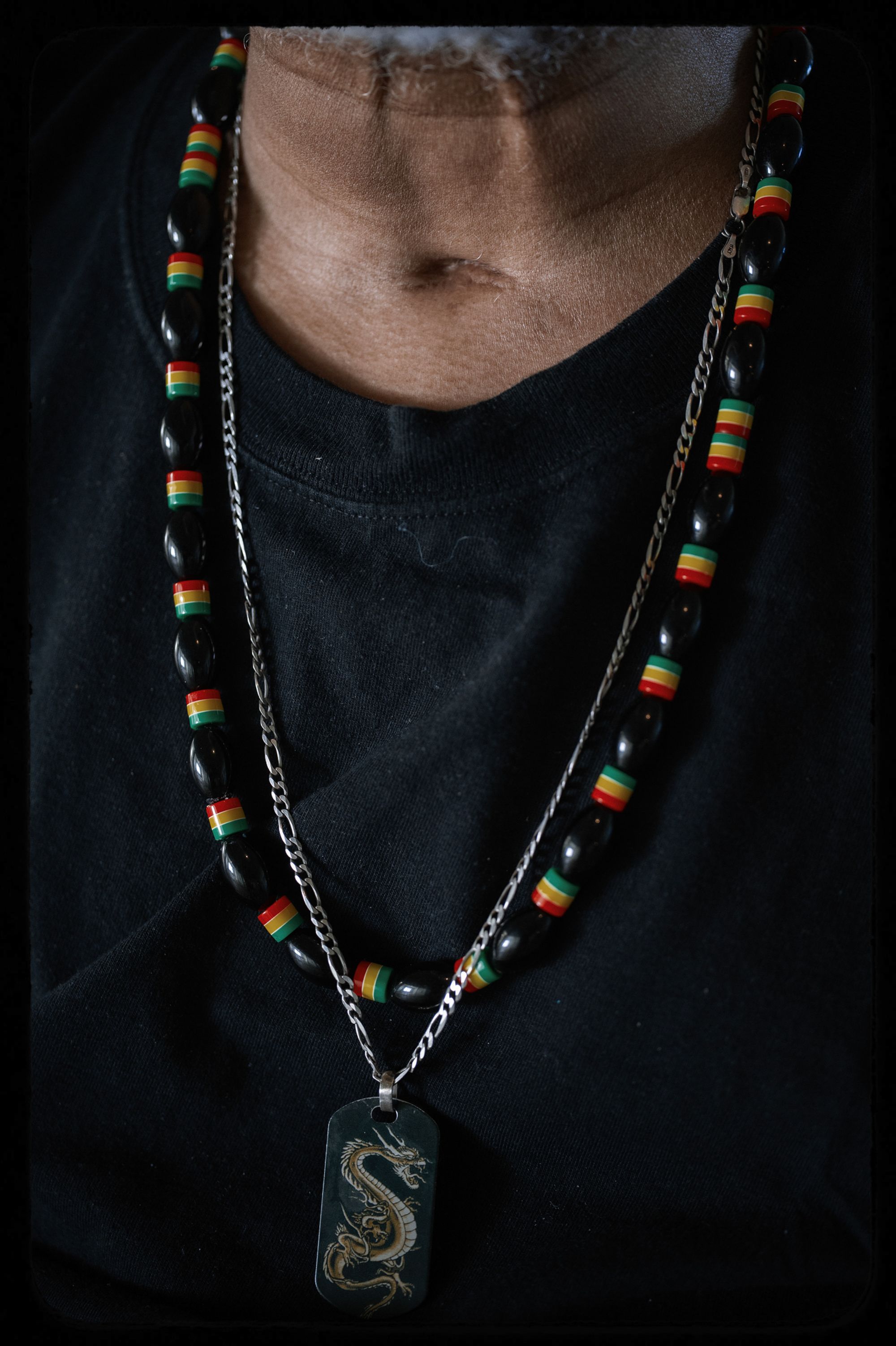 A photo shows a close-up of Sitawa Jamaa’s neck, around which he is wearing two necklaces. One is made up of beads, and the other is a chain with a pendant that has a dragon on it. 
