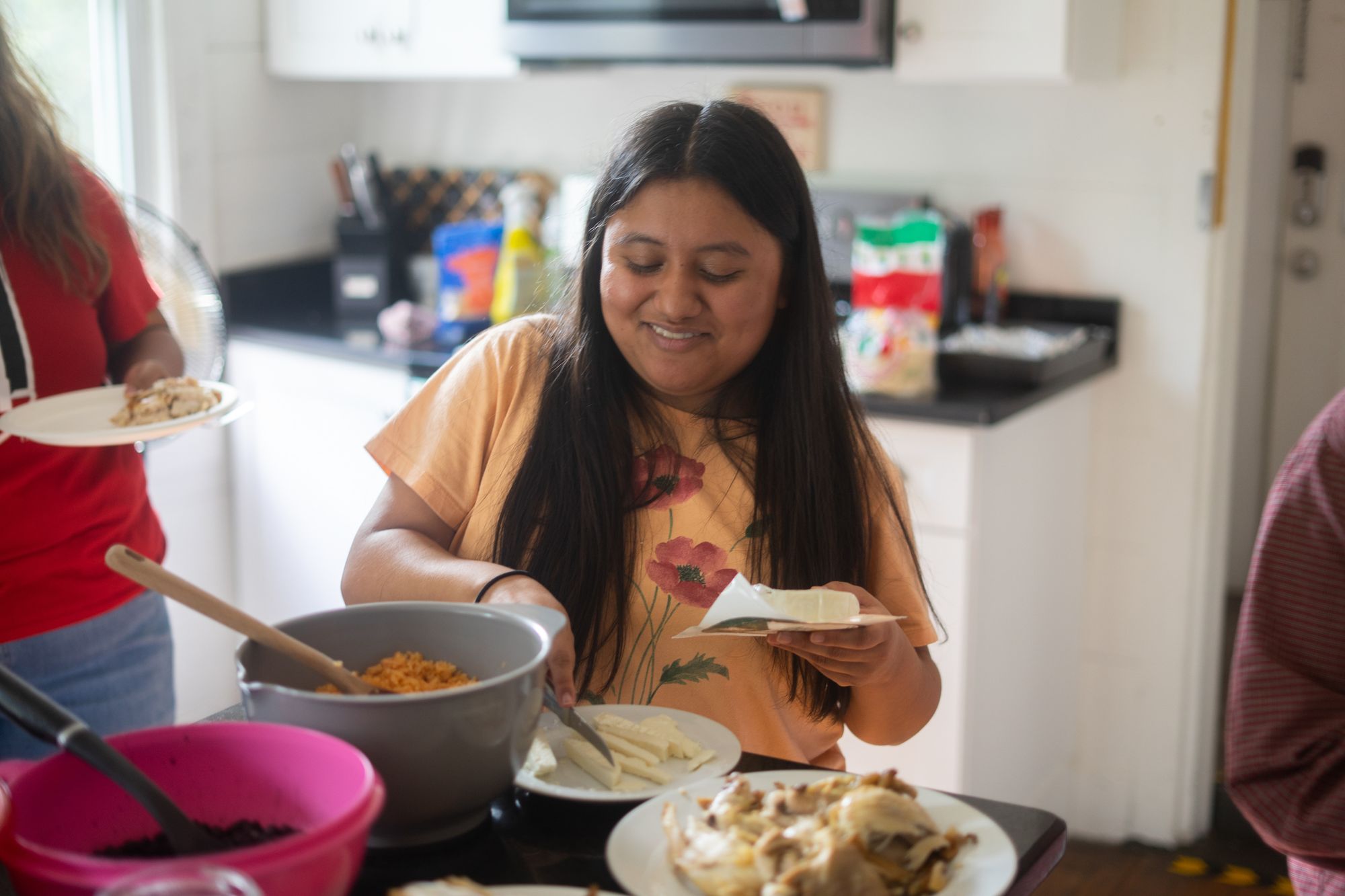Una mujer de piel morena clara y cabello oscuro y largo viste una blusa color durazno con flores. Está sentada en una cocina cortando queso. En el mostrador frente a ella hay un plato de pollo y un tazón de arroz, y una mujer con una camiseta roja está de pie a su derecha.