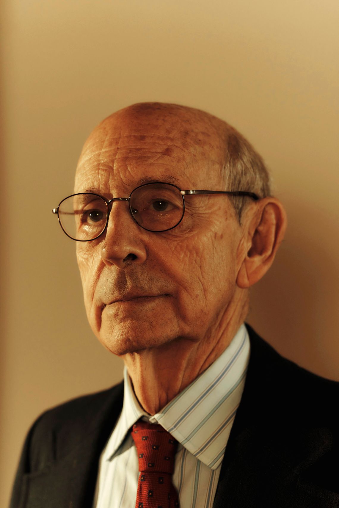 A headshot of an older White man wearing glasses, a black suit, striped shirt and red tie. 