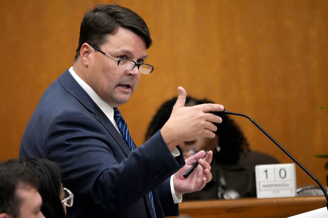 A White man, wearing glasses and a navy suit, speaks into a courtroom microphone.