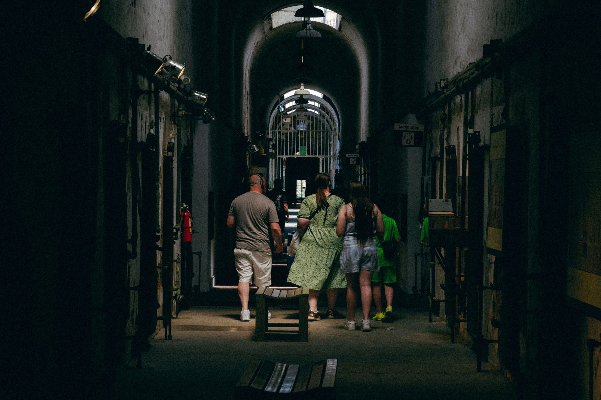 A group of people walk through a dark hallway with prison cells on either side.