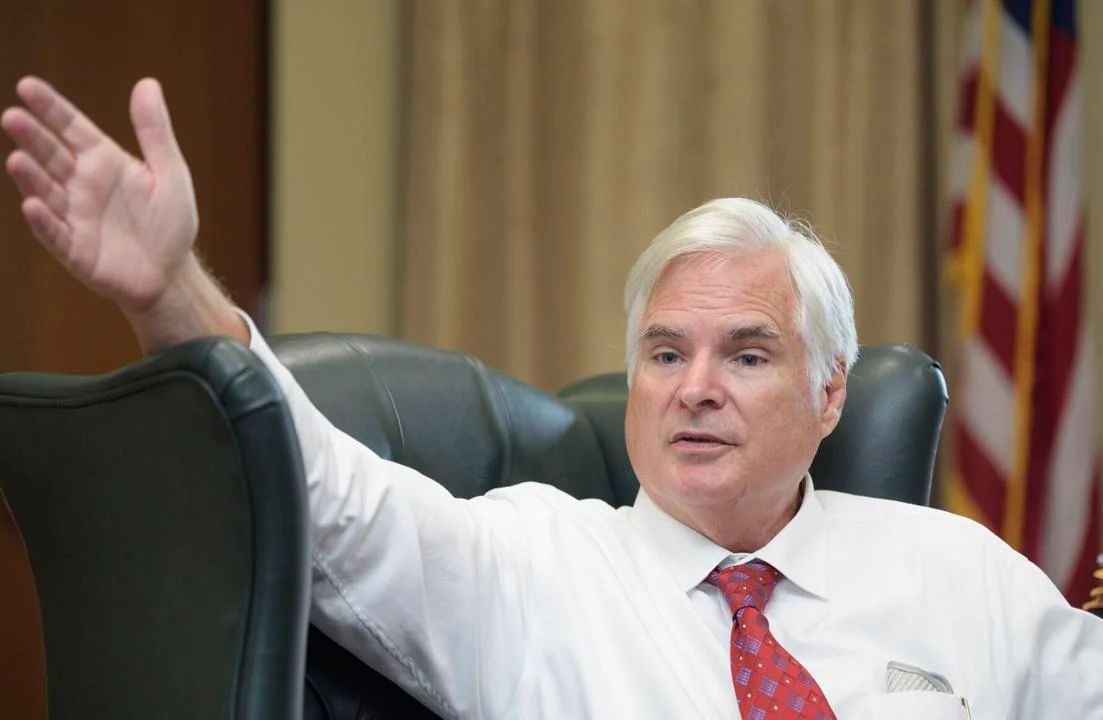 A White man with short white hair, wearing a white shirt and red tie, sits and holds his hand out towards the camera. 