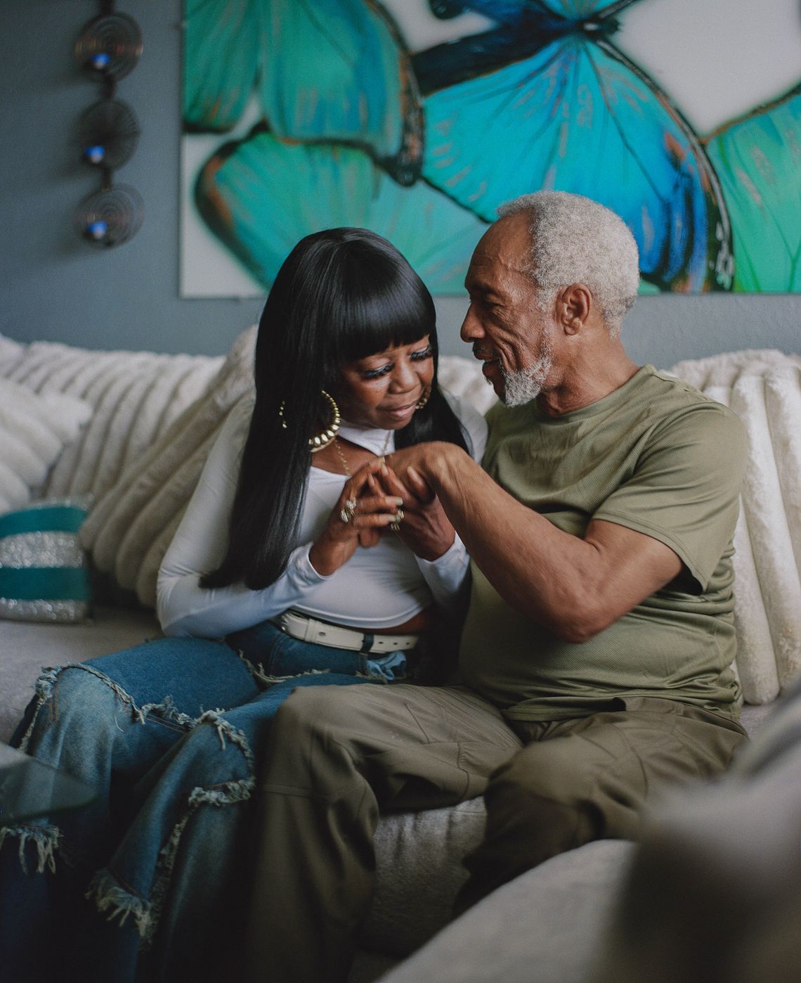 Armour, a Black woman wearing dark jeans and a white long sleeved shirt with a chest cutout, and Moses, a Black man wearing an olive green T-shirt and pants, sit on a couch holding hands. A painting of blue butterflies is visible in the background.  