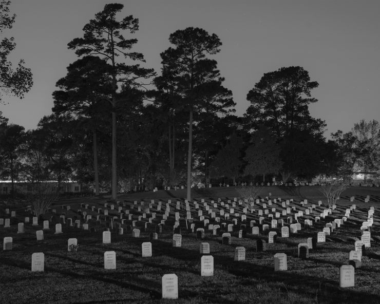 A black-and-white photo shows dozens of gravestones in a cemetery at night. In the background are a number of tall trees.