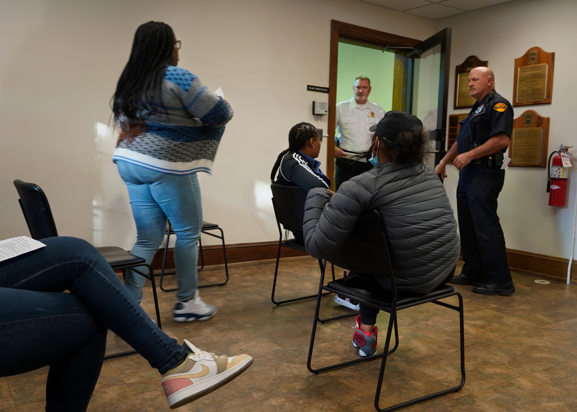 A Black woman stands to attend a hearing for a traffic citation in a waiting room.  A few other people sit nearby.  Two officers stand near the door.  