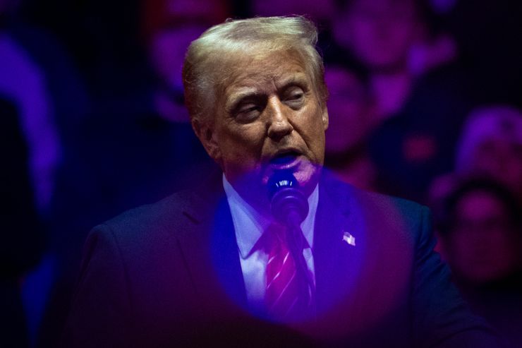 A photograph of President Trump, a White man in a suit and tie, speaking at a podium behind pink and blue orbs of light. 