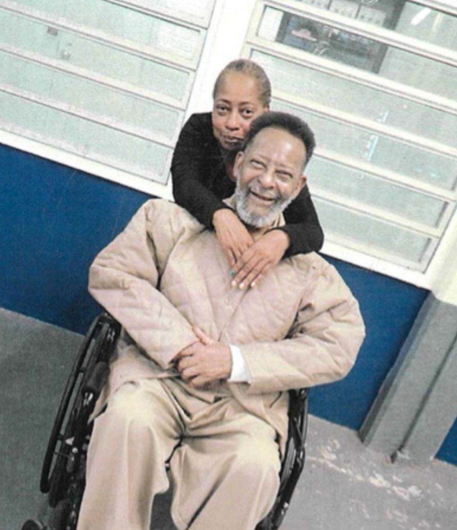 An older Black man with a gray beard and dressed in a khaki-colored jacket and pants sits in a wheelchair with his hands clasped together in front of him.  His daughter, a Black woman with closely cropped salt-and-pepper hair and wearing a dark colored shirt, leans over his shoulders from behind to hug him around his neck. Behind them are a set of horizontal panel windows. 
