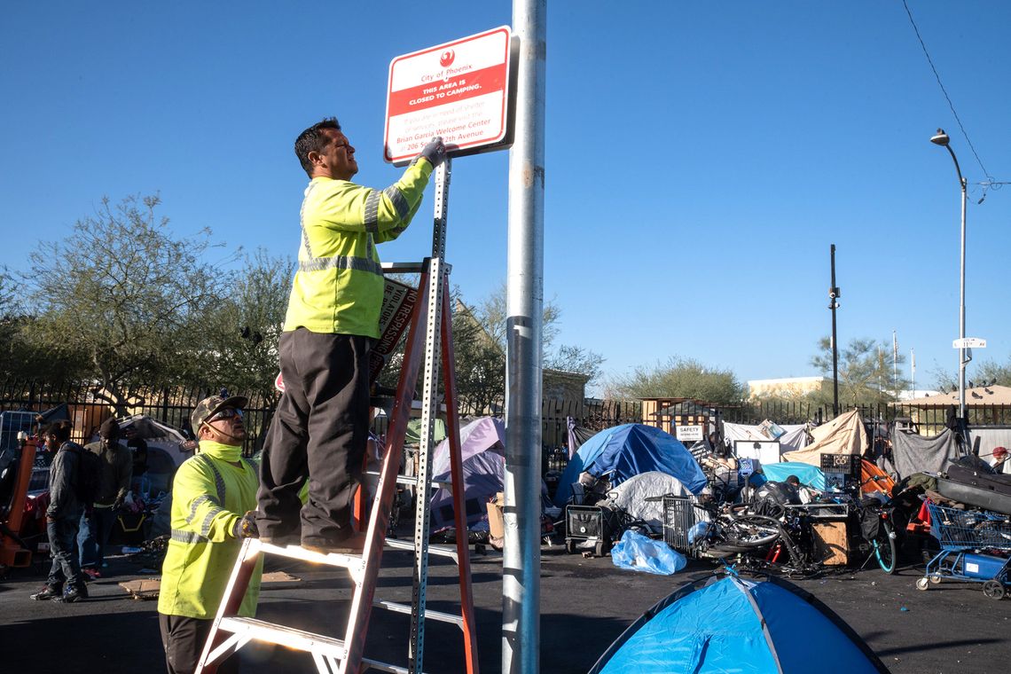 A city worker wearing a neon safety jacket and dark gray pants stands on a ladder while installing a sign on a pole that reads “City of Phoenix: This area is closed to camping.” A second city worker holds the ladder. Tents and other items are lined up on the sidewalk on the street in the background. 