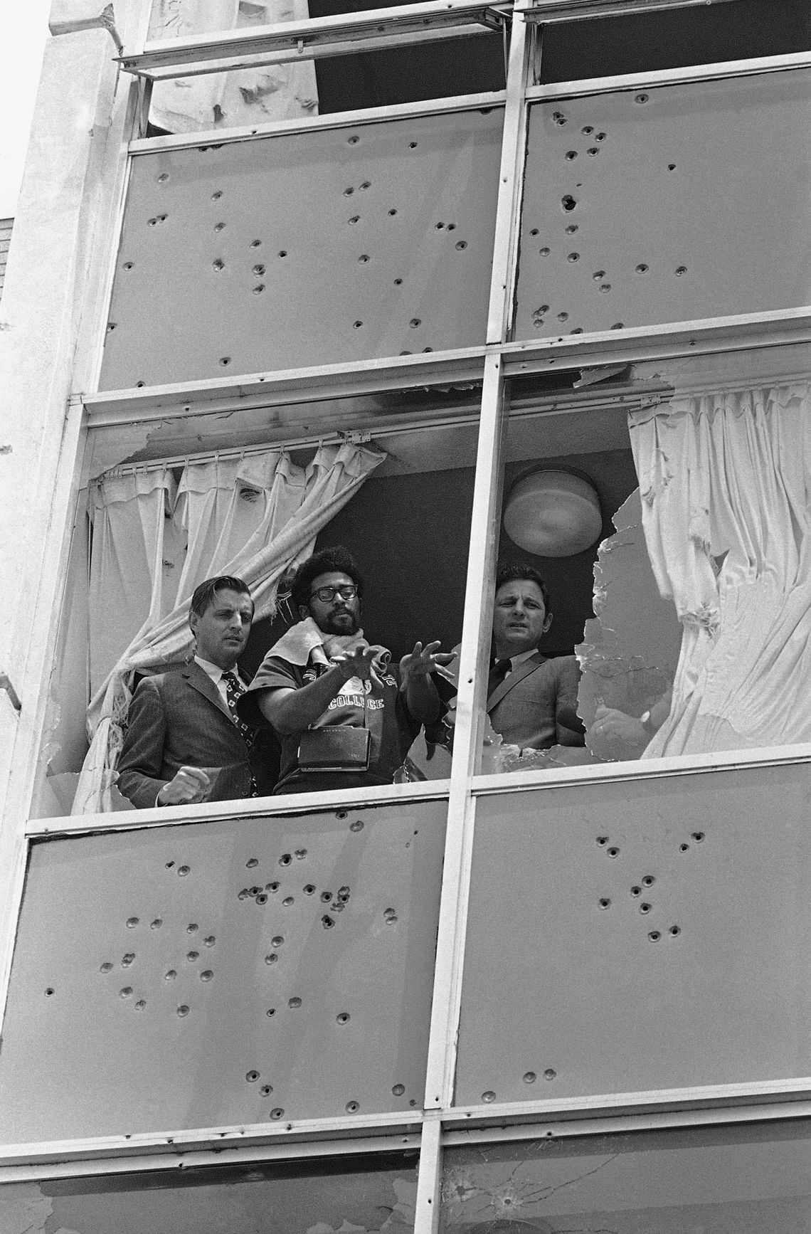 A black-and-white photograph shows two White men in suits looking out a shattered window, while a Black man in a T-shirt gestures downward. The parts of the building around the window are covered in bullet holes. 