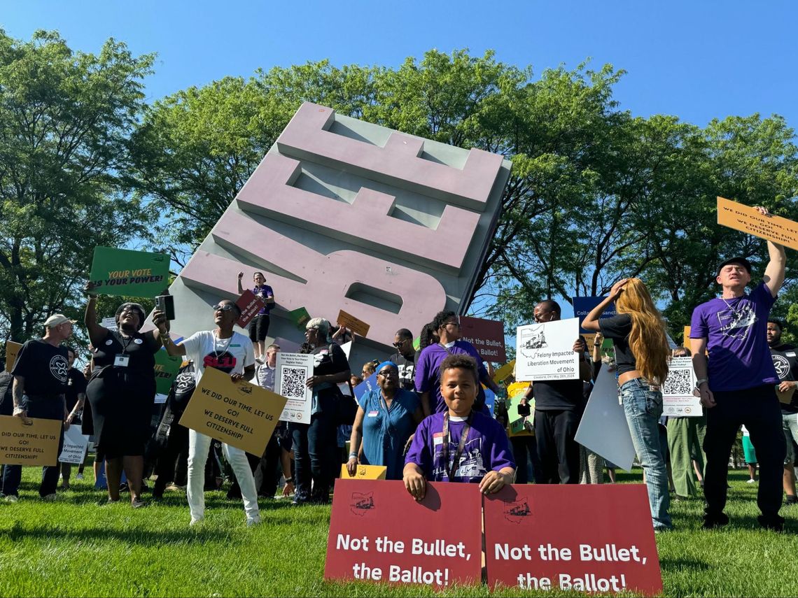 A diverse group of people on a grassy area in downtown Cleveland support Building Freedom Ohio with signs. A child in a purple shirt sits with two signs that say, "Not the bullet, the Ballot!"