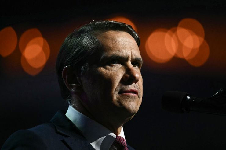 A photo shows the head and shoulders of a man with light-toned skin and short graying hair. He is wearing a suit. Behind him are blurred lights.. 