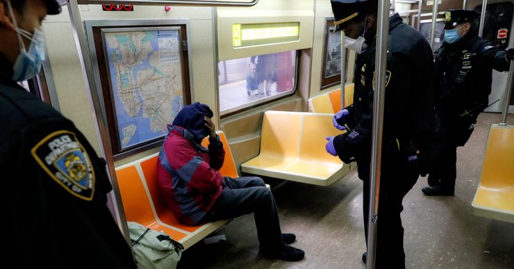 Three standing police officers surround a sitting passenger in a subway car. The passenger sits hunched over, with their hand on their head. 