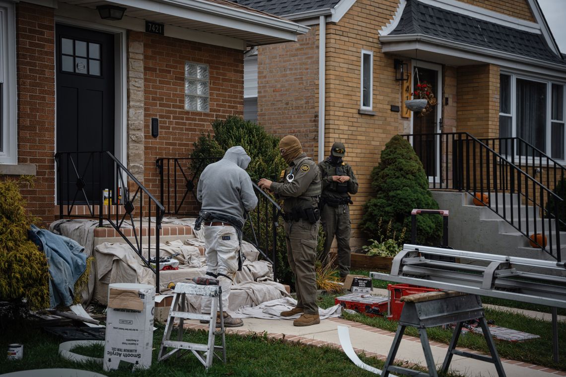 A photo shows two masked federal agents with a person wearing work clothes in front of two brick homes. Tarps, tools and other supplies are around the home in the foreground. 