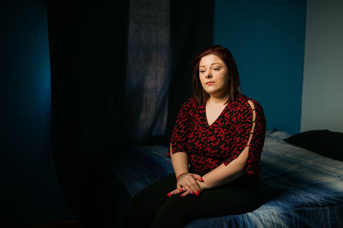 Rebecca McDonald, a light-skinned woman with straight brown hair, wears a black and red patterned top while sitting in her son's bedroom, which has blue walls and a blue and white blanket on the bed. 