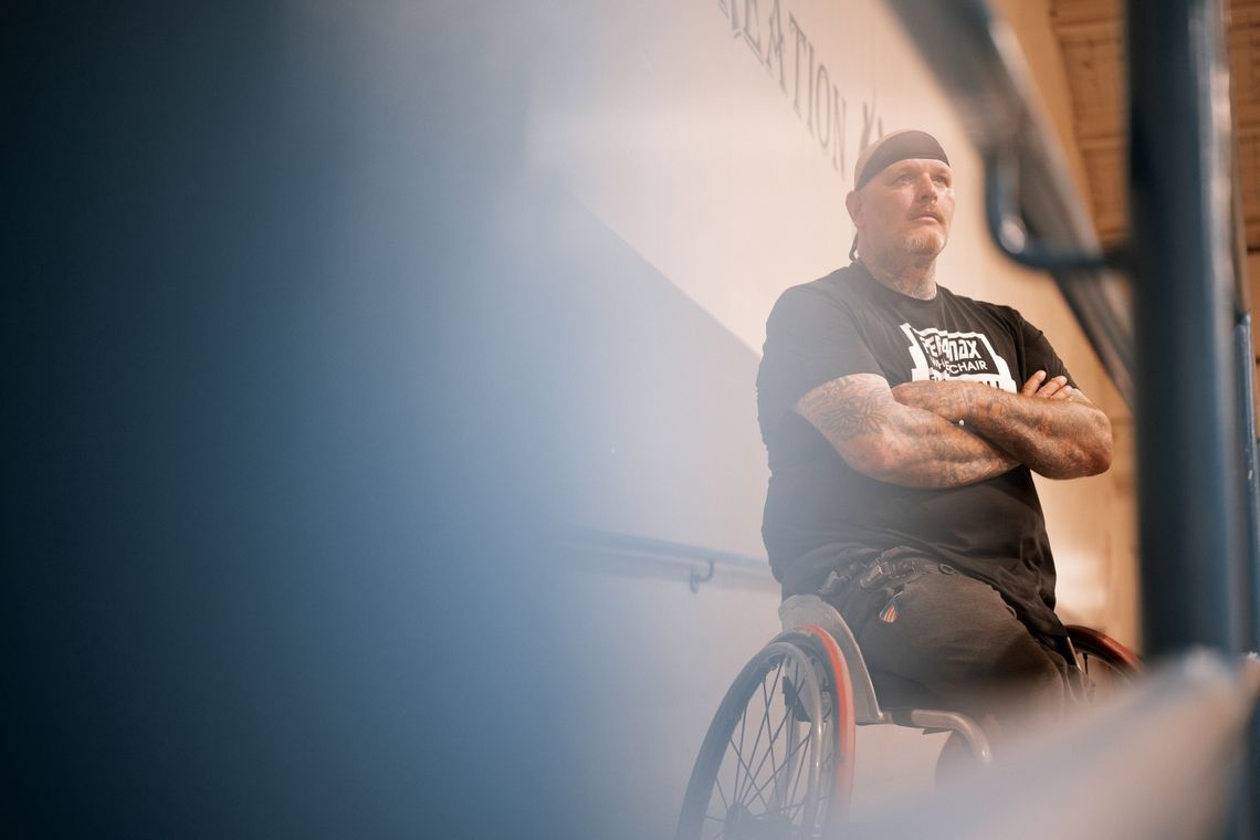 A photo shows a White man wearing a black T-shirt in a wheelchair, wearing a sweatband around his head and with his arms folded. The camera is focused on him and the left side of the image is out of focus. 