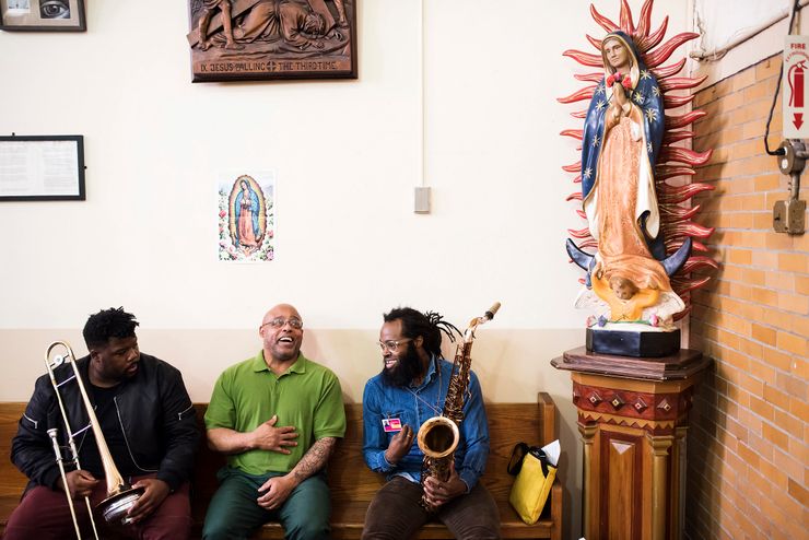 Three Black men sit in a chapel, next to a statue of Our Lady of Guadalupe. The man sitting in the middle wears glasses and a lime green polo over his dark green prison uniform. The two men sitting on either side of him hold a trombone and a saxophone, wearing jackets and slacks. 