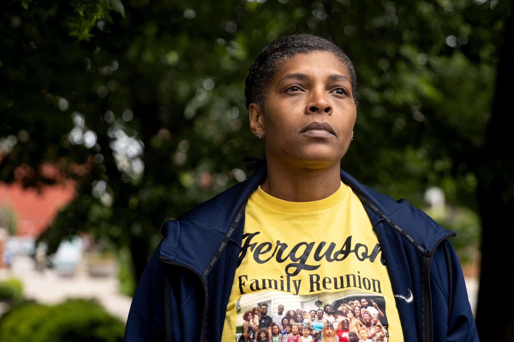 A photo of a middle-aged Black woman with her hair up posing for a portrait outdoors. She is wearing a yellow shirt with a family photo under the words “Ferguson Family Reunion.”