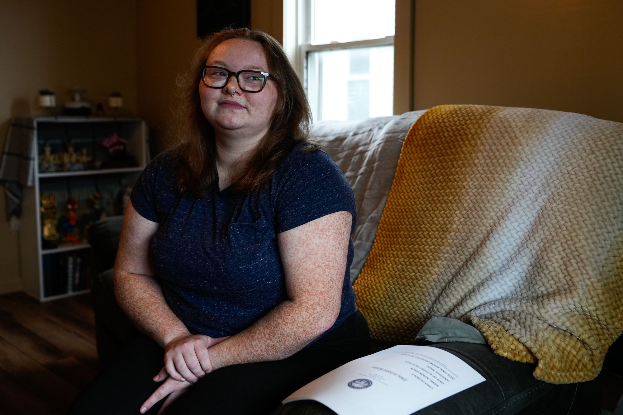 A photo shows Katherine Dixon, a White woman wearing glasses and a dark blue T-shirt, sitting on a couch. 