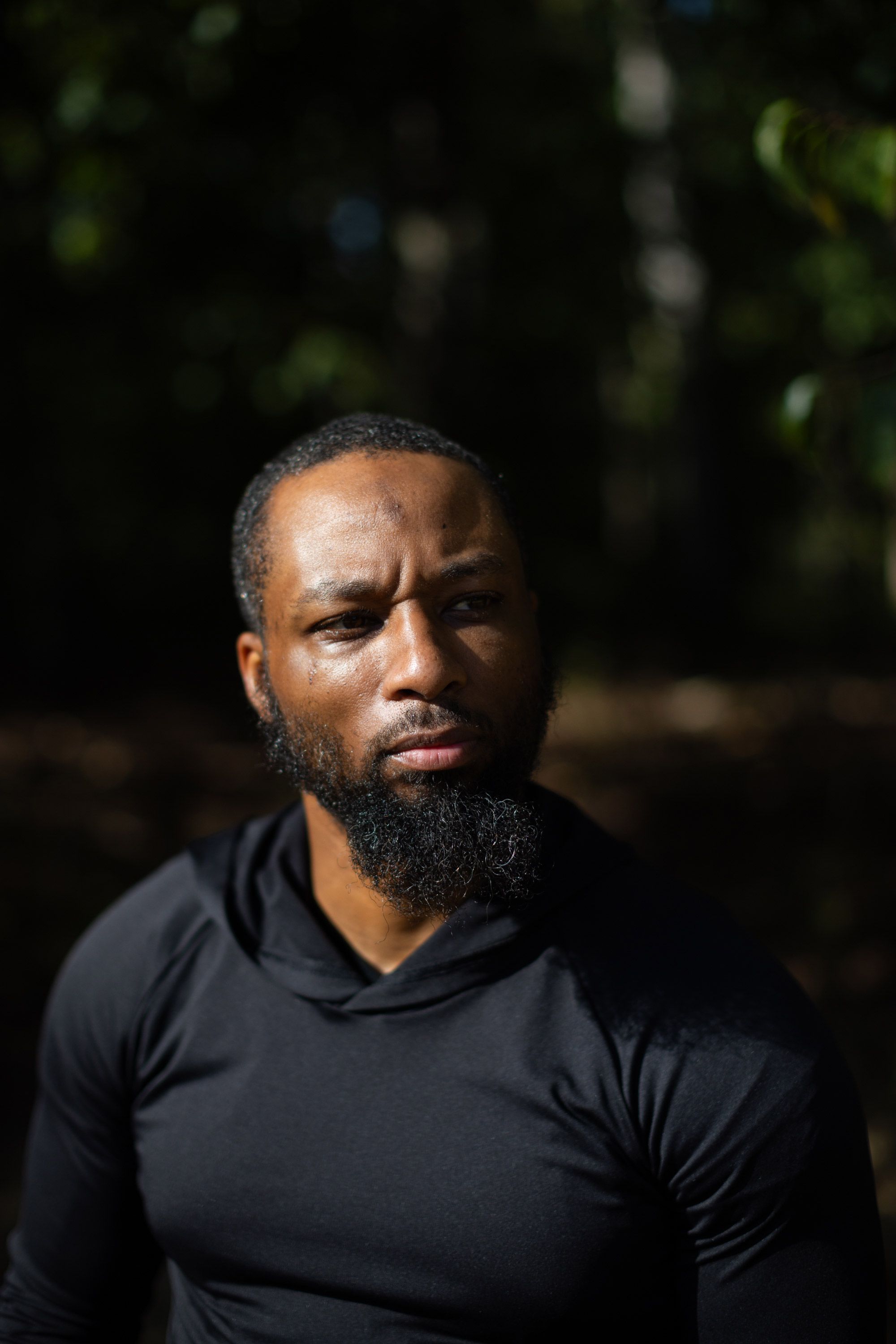 A photo of a Black man with a beard and a black top. The man is looking to the side with a furrowed expression.