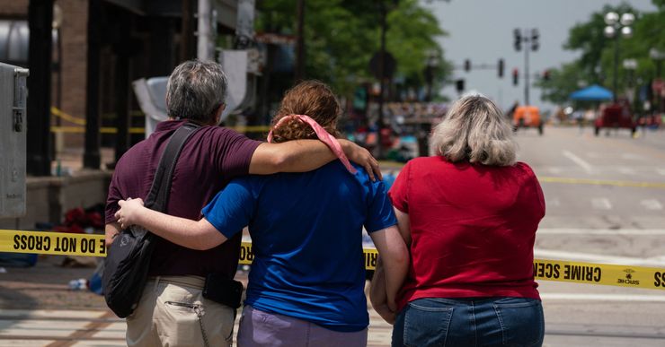 A man with gray hair, wearing a plum-colored t-shirt, has his arm around a woman with reddish-brown hair, wearing a blue t-shirt.  She is holding hands with a woman with blonde hair, wearing a red t-shirt.  They all stand in front of police tape, looking down the street.  