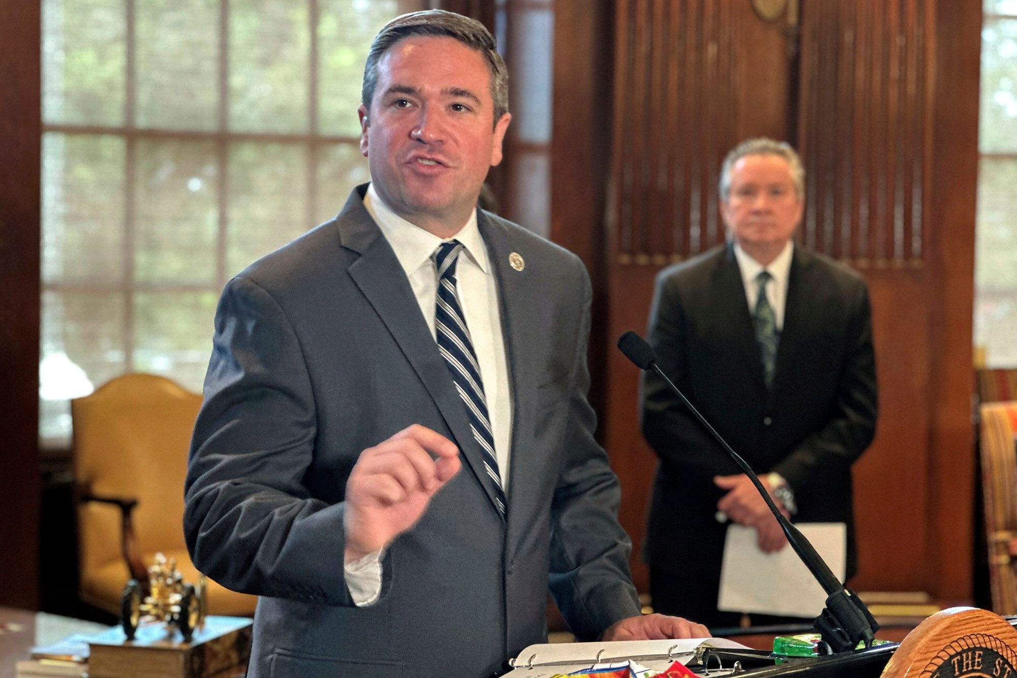 Missouri Attorney General Andrew Bailey, a White man wearing a gray suit, speaks into a mic at a podium. A White man, wearing a black suit and holding a piece of paper, stands in the background. 