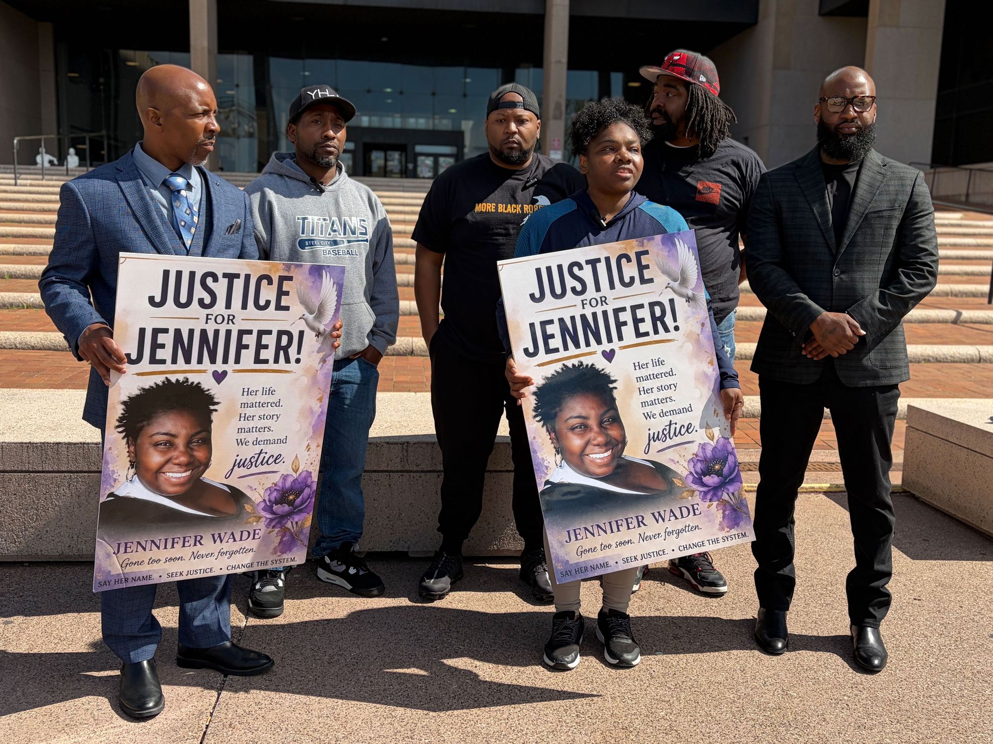 A photo of a group of six middle-aged Black people in front of a building. In the front row, two people in blue, one of them a lawyer in a suit, hold signs that read, "Justice for Jennifer!" with a photo of a smiling Black woman.  