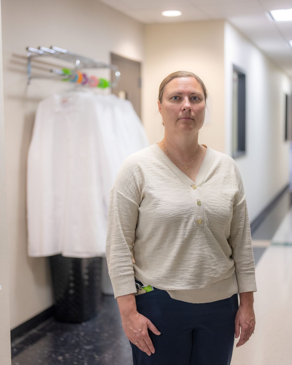A photo shows a White woman wearing a cream-colored shirt and black pants. Lab coats are hung on a rack behind her, and part of a hallway is visible. 