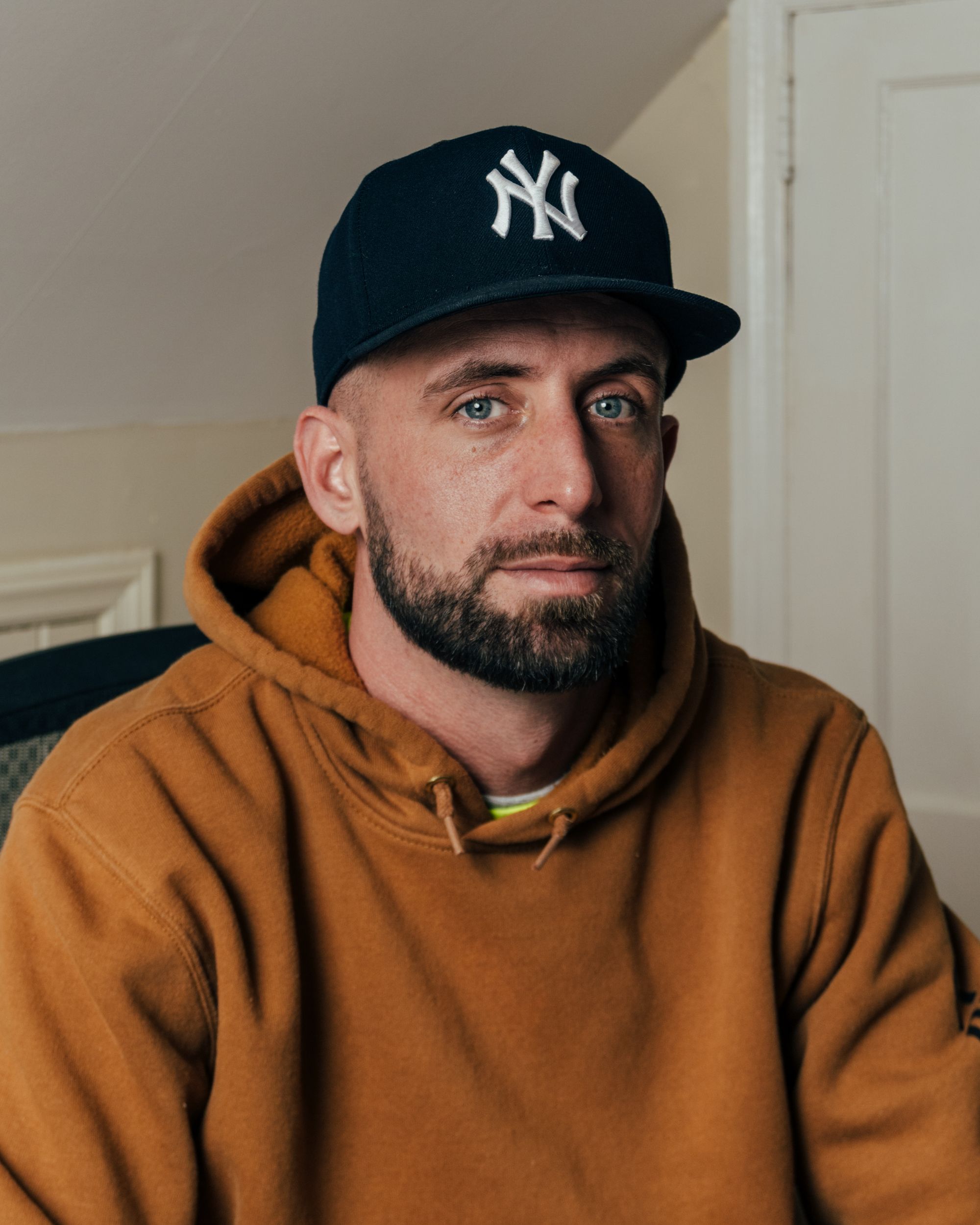 Nick Magalios, a White man, wears a black New York Yankees baseball cap and a burnt orange hooded sweatshirt. 